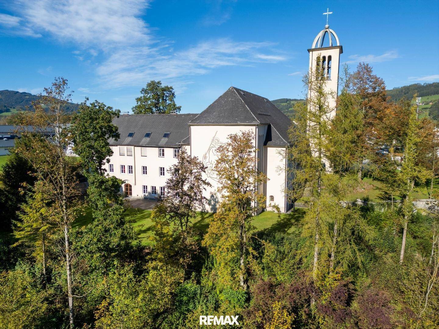 Luftaufnahme eines großen Kirchengebäudes mit Glockenturm, umgeben von Bäumen und Grünflächen.