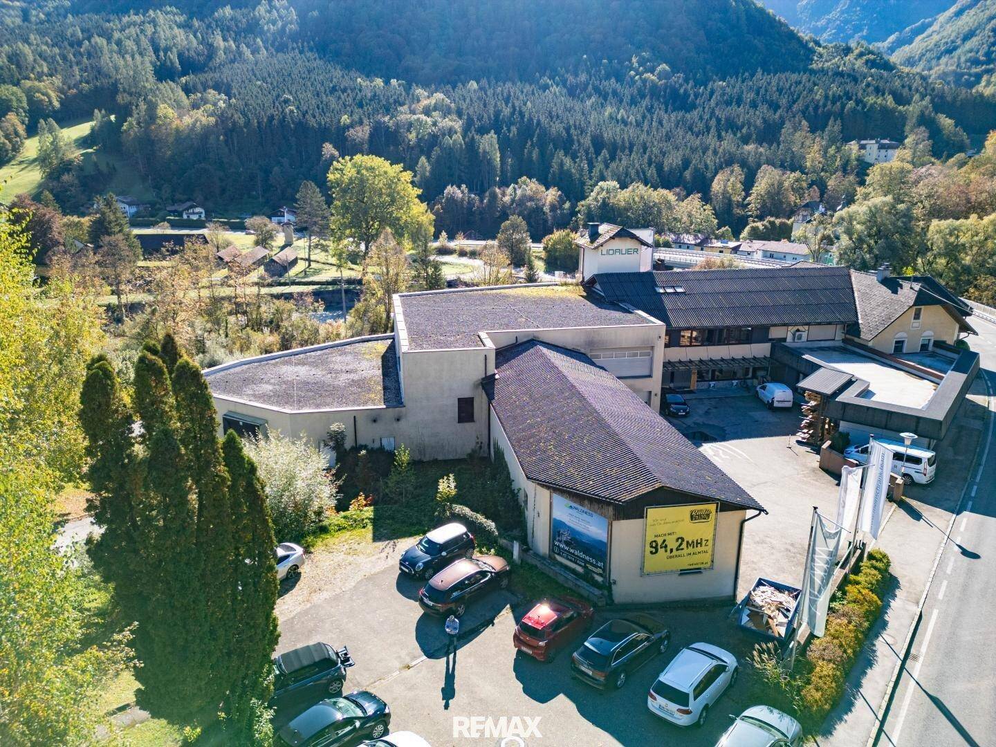 Luftaufnahme des Gebäudekomplexes, umgeben von grünen Wäldern und Bergen, mit Parkplätzen vor dem Eingang.