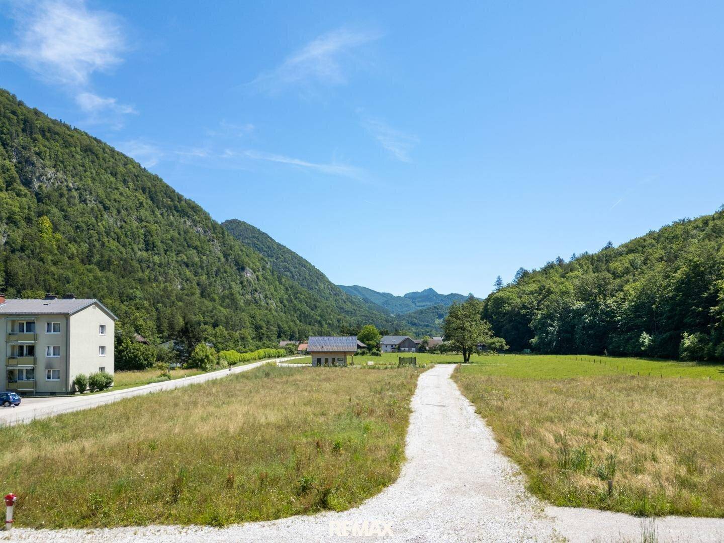 Weitläufiges Wiesengrundstück mit Schotterweg, umgeben von grünen Bergen und vereinzelten Häusern.