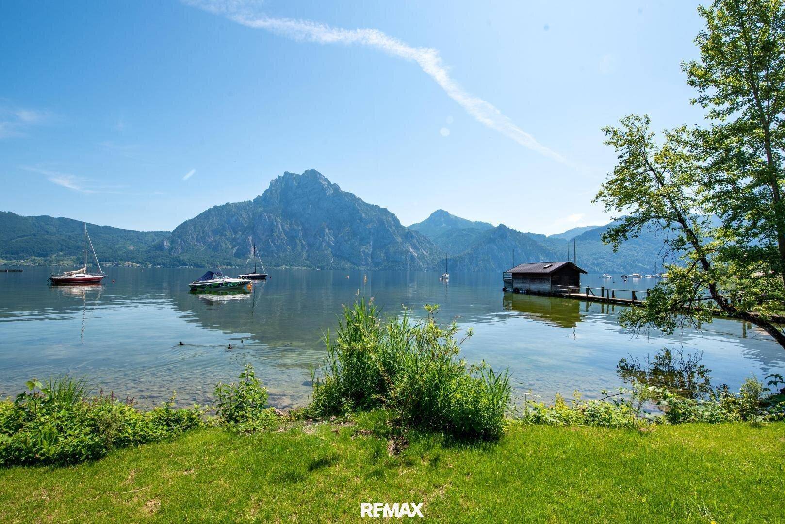 Panoramablick auf den See mit Bergen und einem Bootshaus unter blauem Himmel.