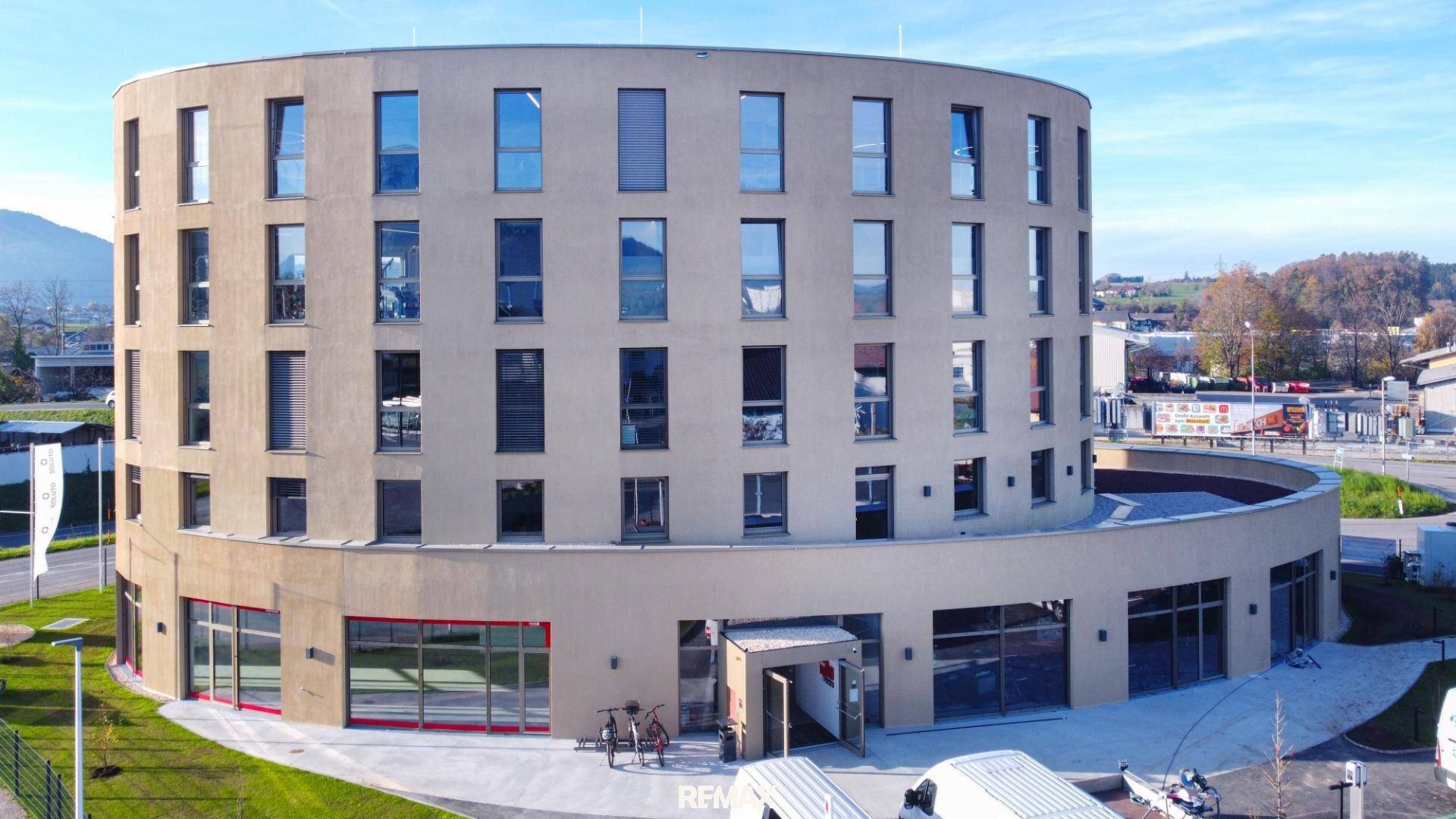 Modernes, rundes Gebäude mit beiger Fassade, großen Fenstern und Blick auf die umliegende Landschaft und Berge.