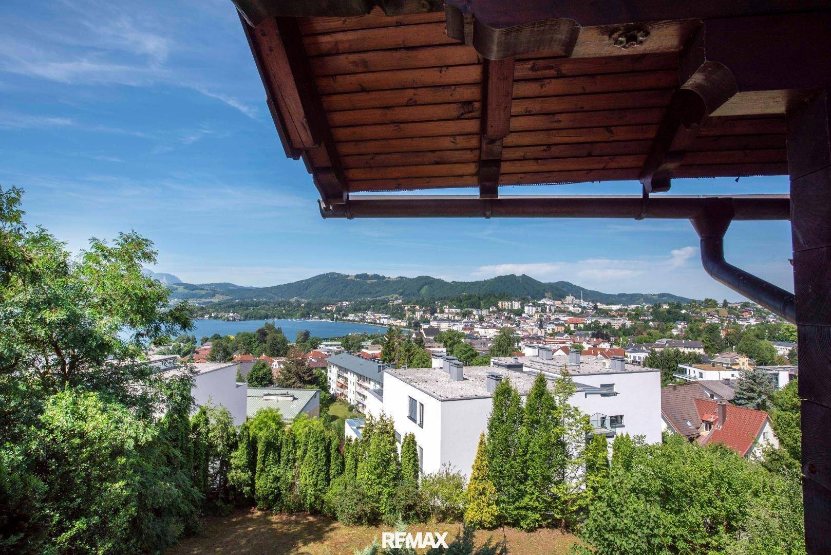 Panoramablick von einem Balkon auf einen See, die Stadt und die umliegenden Berge unter blauem Himmel.