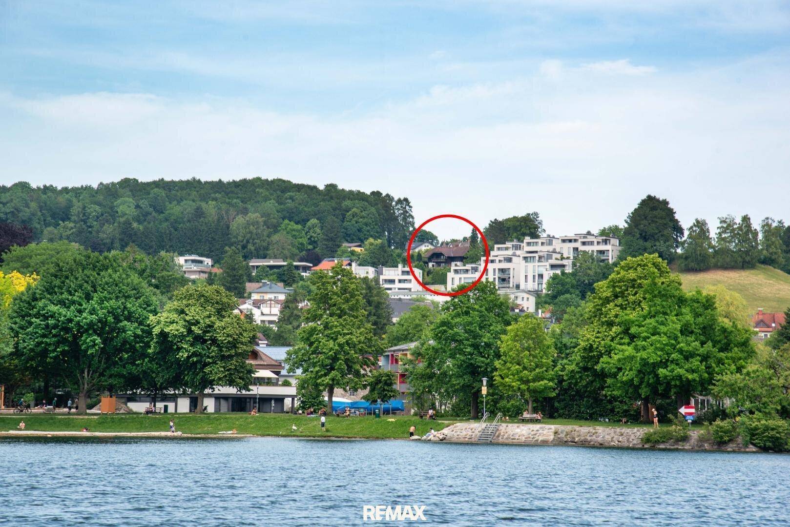 Panoramablick über den See mit bewaldeten Hügeln und der Stadt im Hintergrund, das Objekt ist rot markiert.