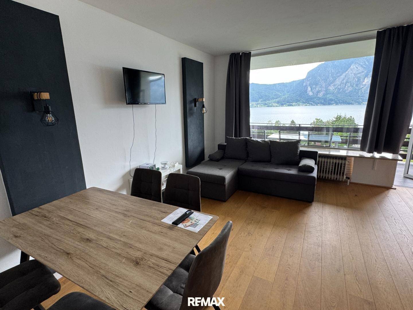 Helles Wohnzimmer mit Holzboden und großem Fenster mit Blick auf See und Berge.