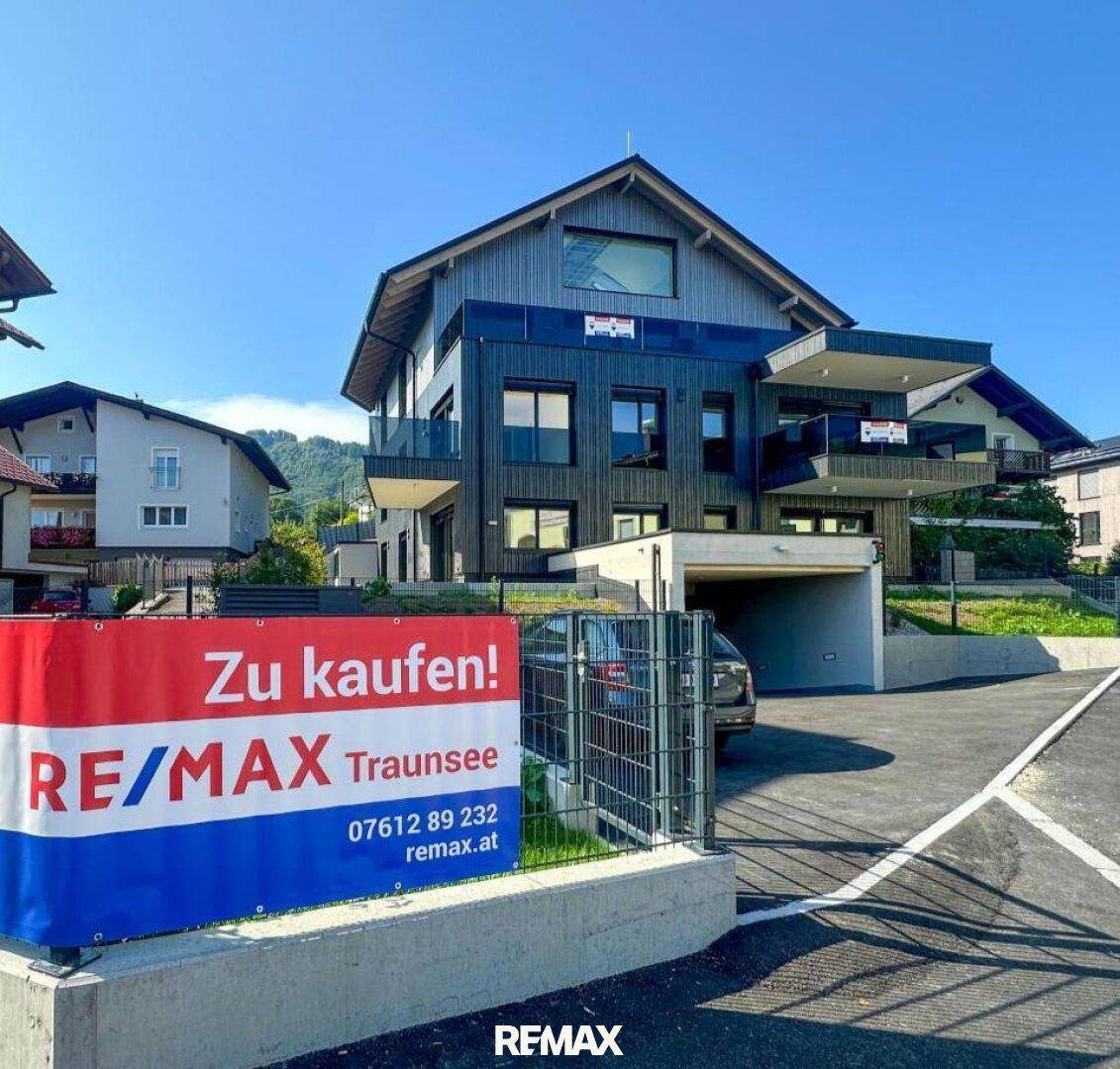 Modernes Mehrfamilienhaus mit dunkler Fassade und Balkonen, umgeben von Grünflächen und einer Verkaufsanzeige.