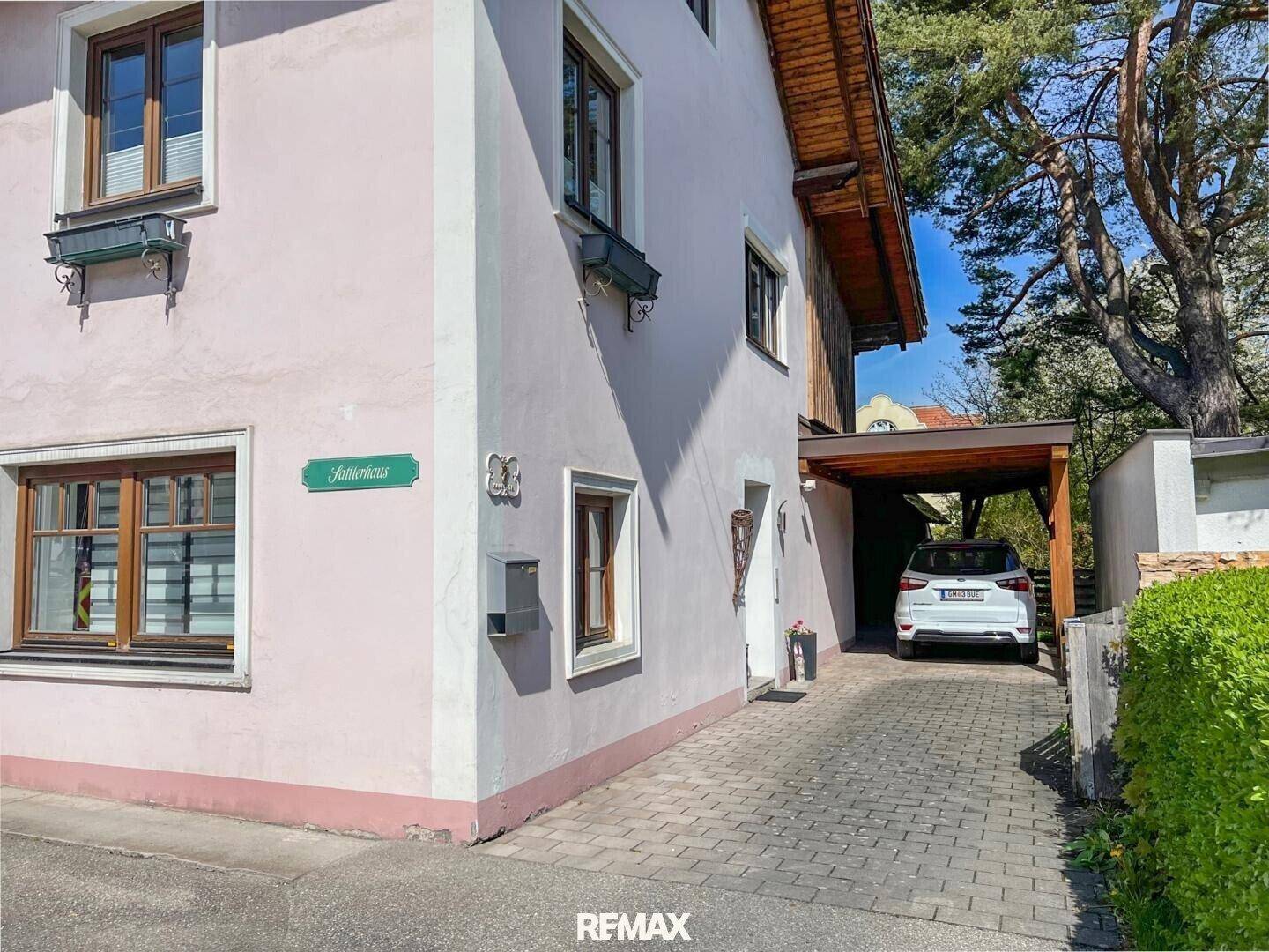 Außenansicht eines gepflegten Einfamilienhauses mit hellrosa Fassade, Holzfenstern, gepflasterter Auffahrt und Carport.
