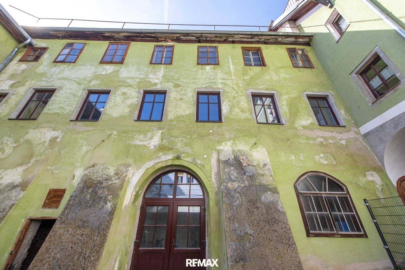 Alte, verwitterte grüne Fassade eines Gebäudes mit vielen Fenstern und einem großen Rundbogeneingang.