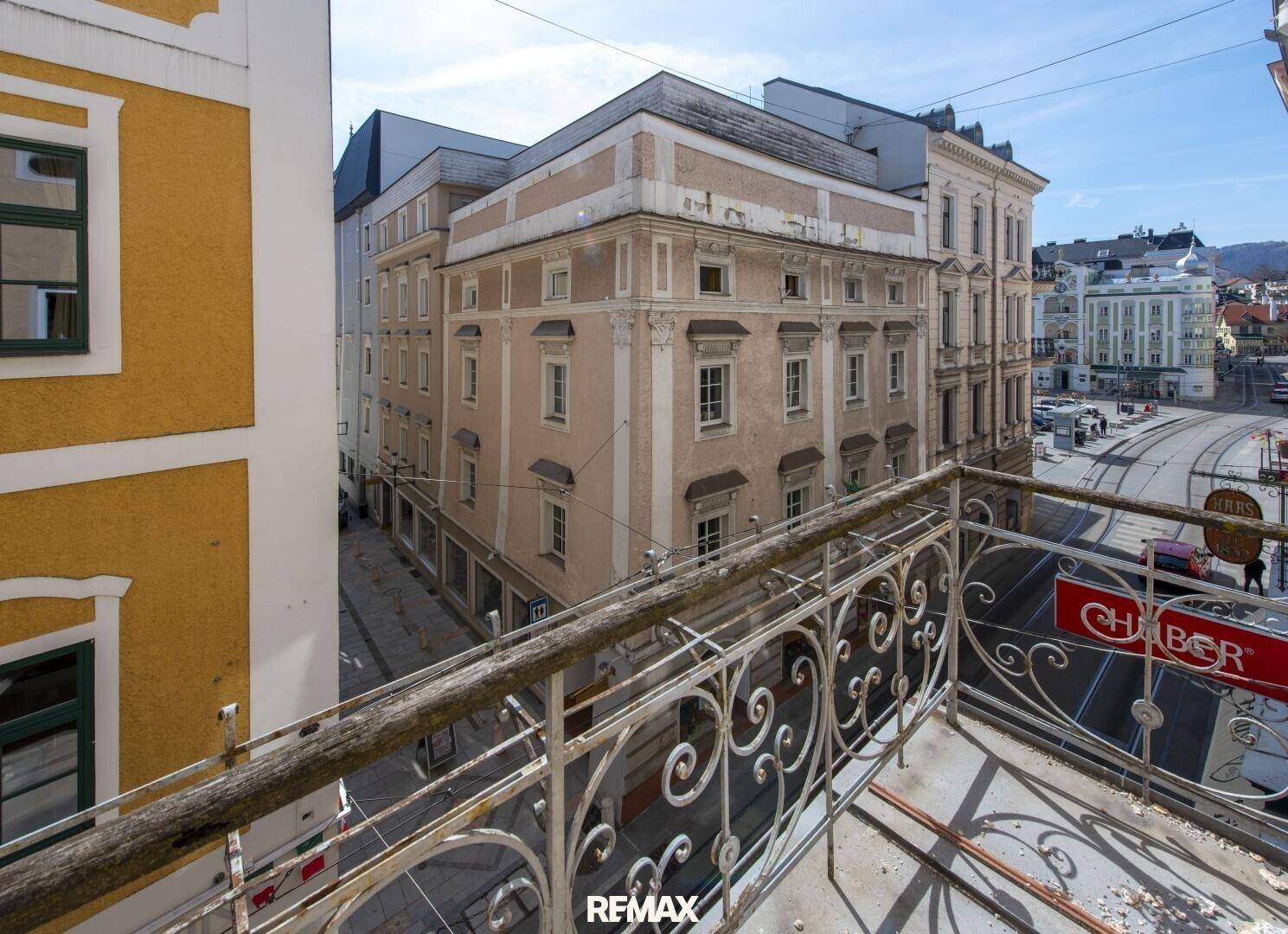 Blick von einem Balkon mit schmiedeeisernem Geländer auf umliegende historische Gebäude und eine belebte Straße.
