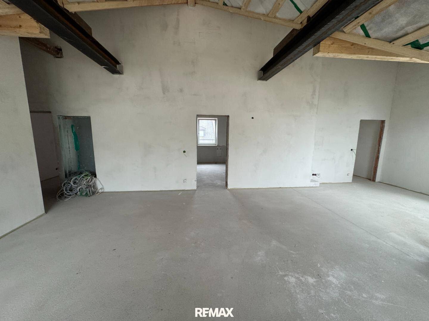 Rohbau eines Innenraums mit Betonboden und sichtbaren Deckenbalken.