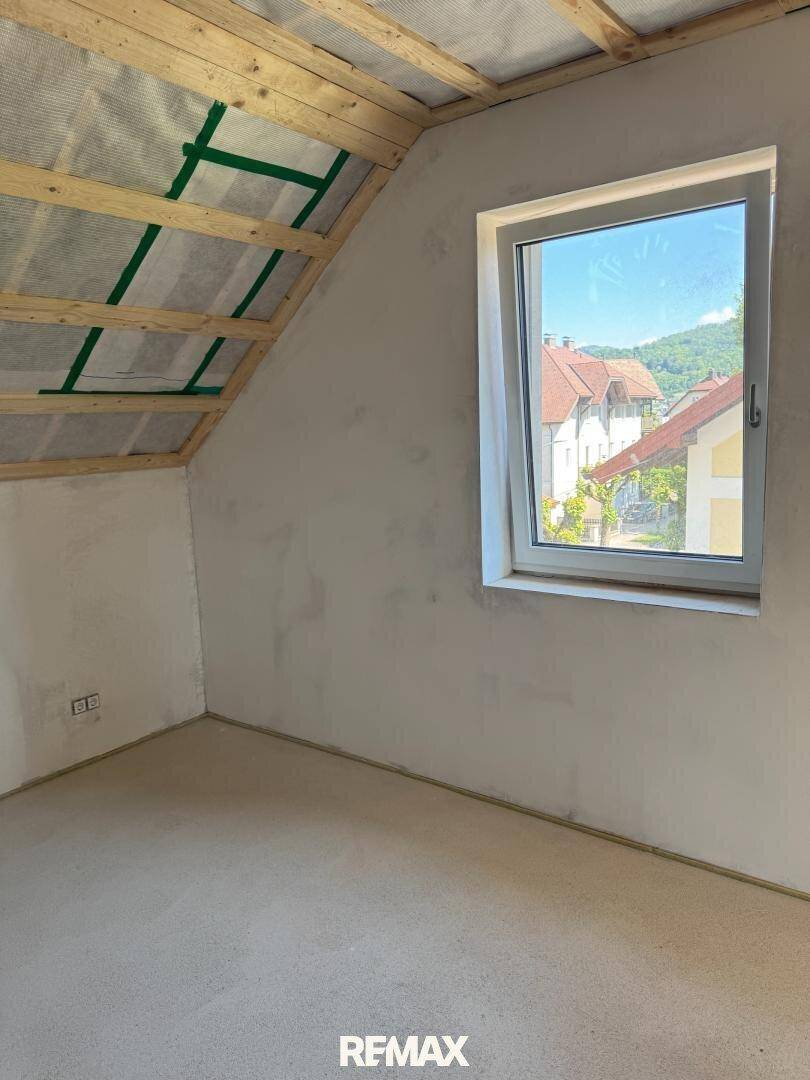 Unfertiger Raum im Dachgeschoss mit sichtbaren Holzbalken und Fensterblick nach draußen.