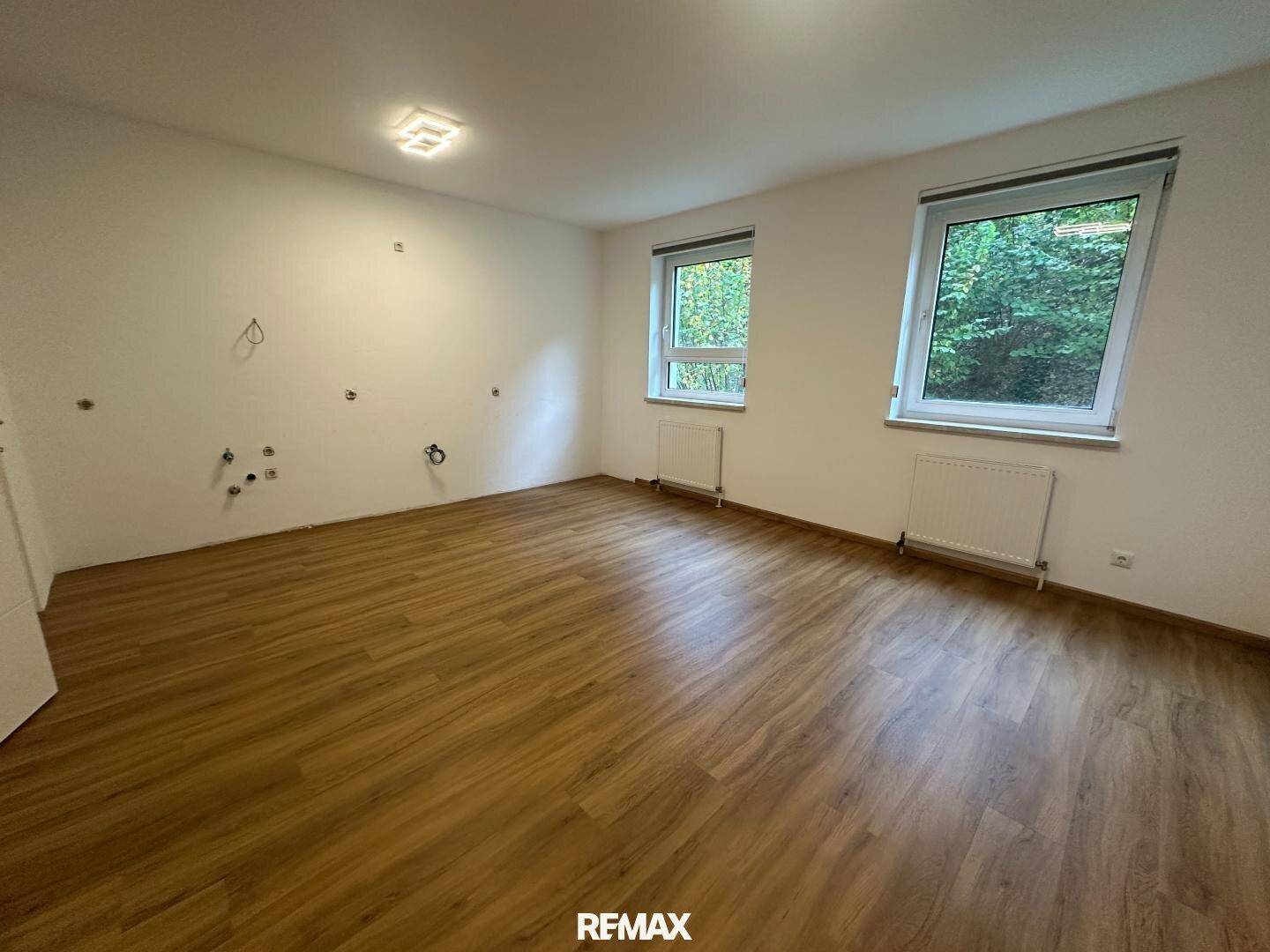 Leerer Raum mit Laminatboden, zwei Fenstern und Anschlüssen für eine Küche.