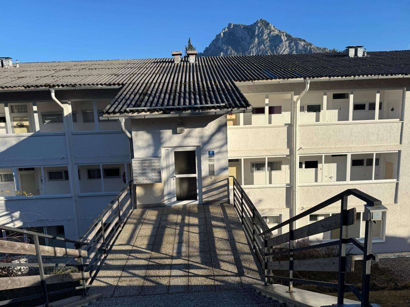 Eingangsbereich eines Mehrfamilienhauses mit Rampe und Treppe, umgeben von einer Berglandschaft.