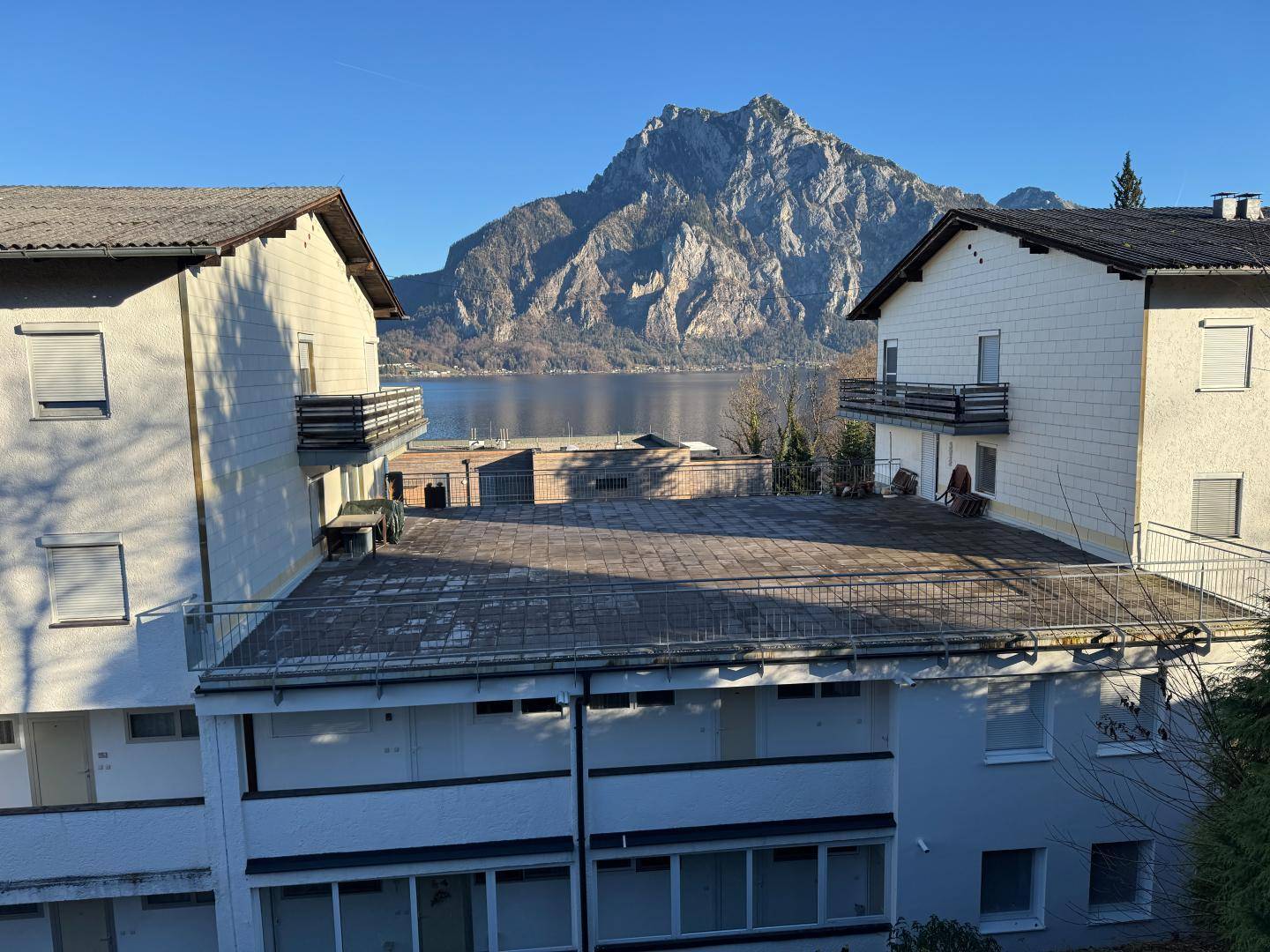 Mehrere Gebäude mit Flachdach und Balkonen, mit einem See und Bergen im Hintergrund unter strahlendem Himmel.