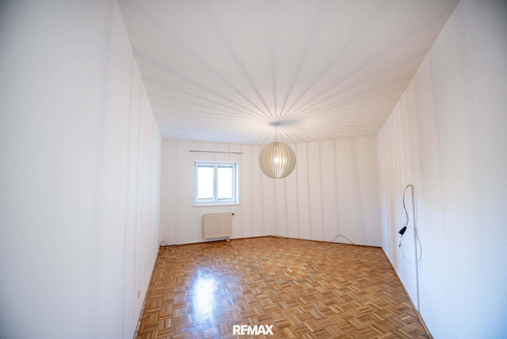 Leeres Zimmer mit Parkettboden, einem Fenster und einer modernen Deckenleuchte.