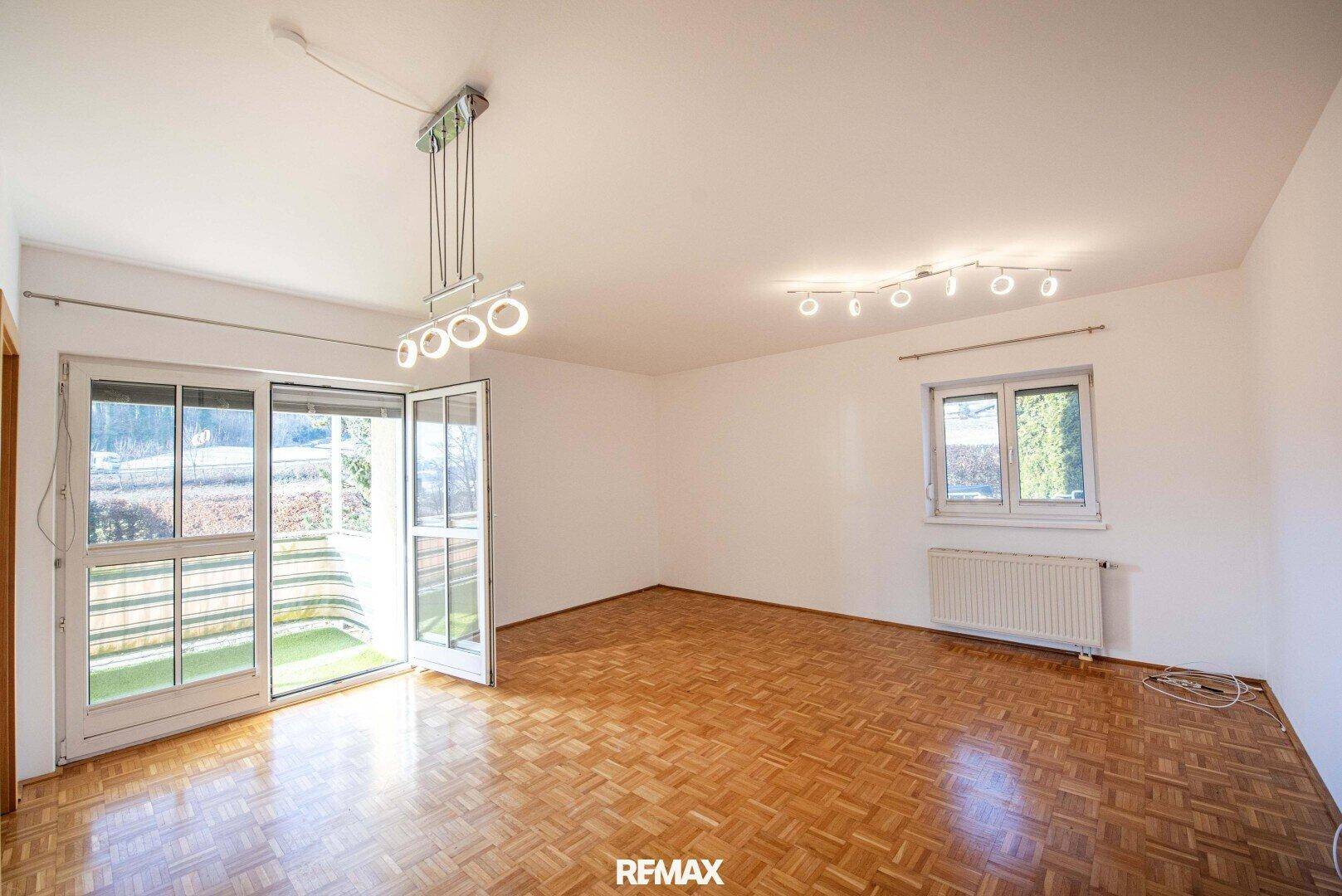 Heller Wohnraum mit Fischgrätparkett, großen Fenstern und Zugang zu einem Balkon.