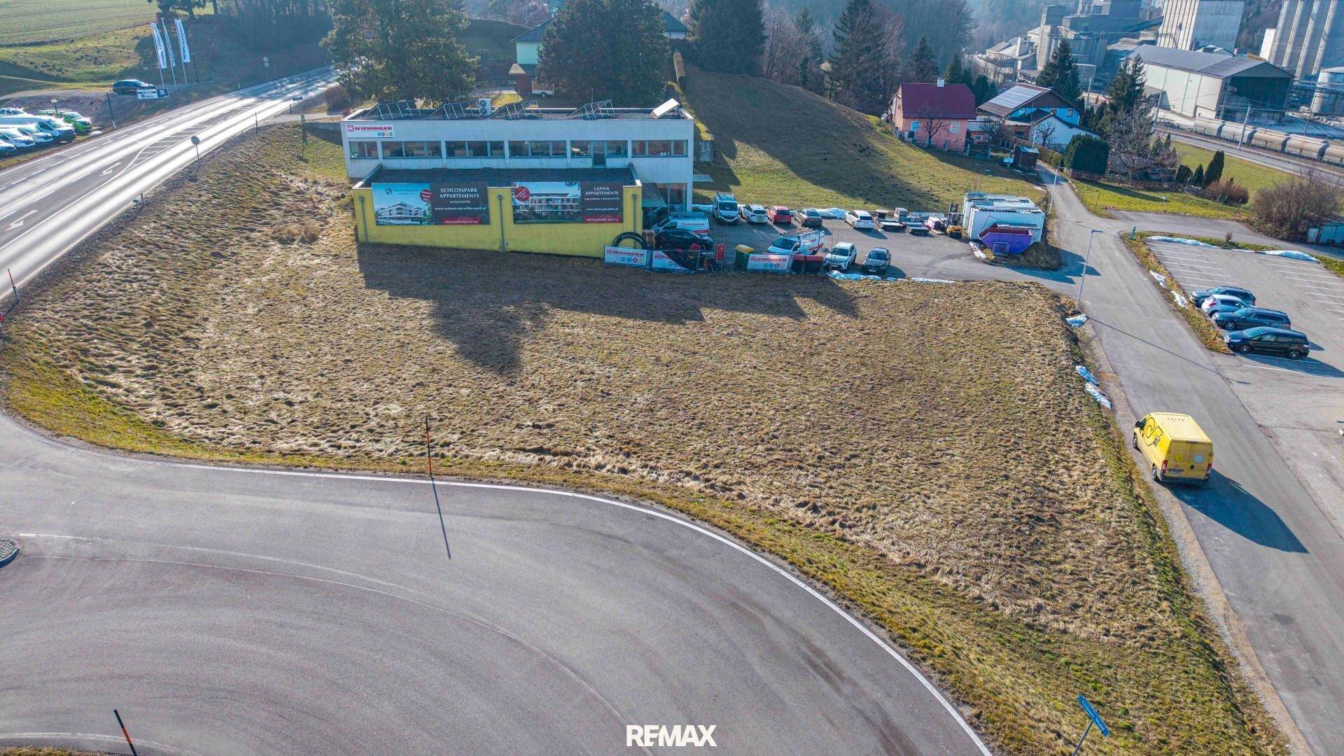 Luftaufnahme eines unbebauten Grundstücks neben einer Straße und einem Gewerbeobjekt mit Parkplätzen.