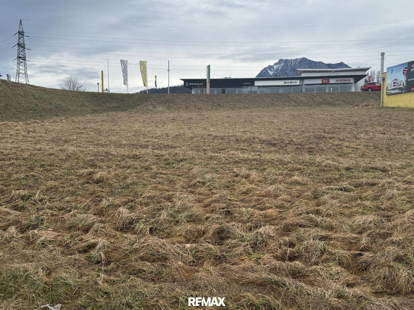 Ungenutztes Grundstück mit Blick auf Gewerbegebäude und Berge unter bewölktem Himmel.