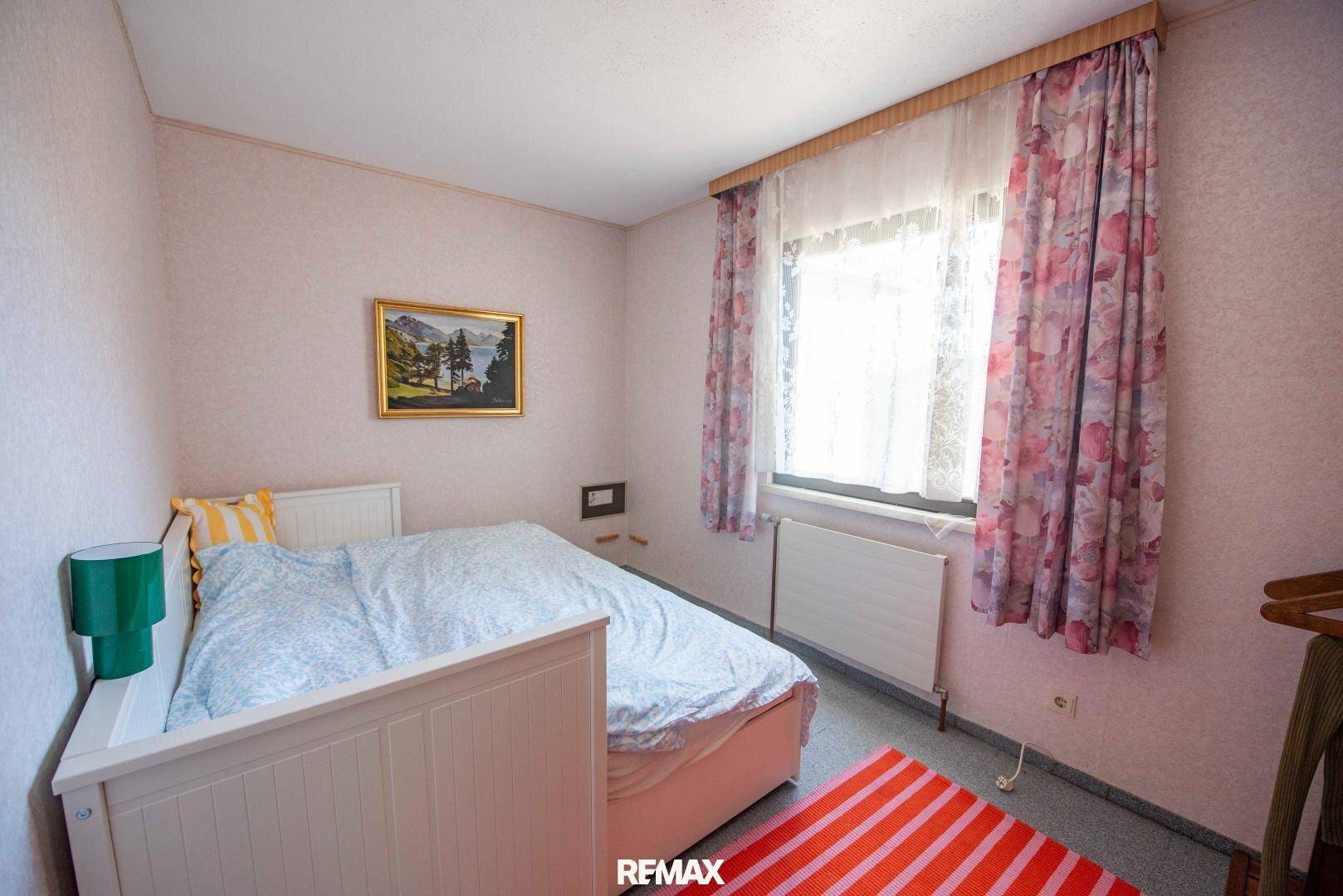 Gemütliches Schlafzimmer mit Einzelbett, floralen Vorhängen und einem Fenster, das Tageslicht hereinlässt.
