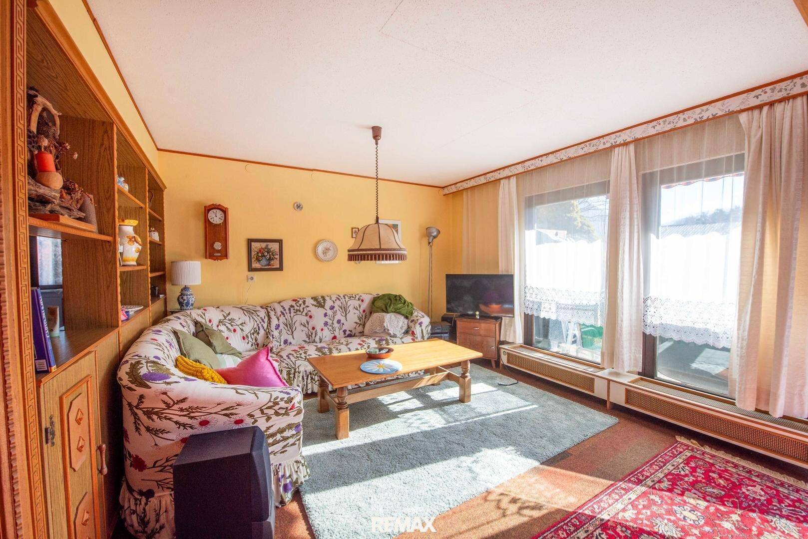 Helles Wohnzimmer mit floralem Sofa, Teppichboden und großen Fenstern mit Blick ins Grüne.