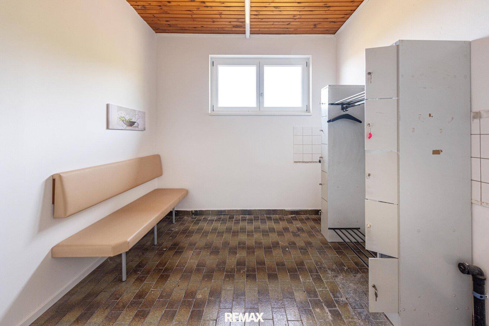 Umkleideraum mit Sitzbank, Spinden, Fenster und Holzdeckenverkleidung, funktional eingerichtet.
