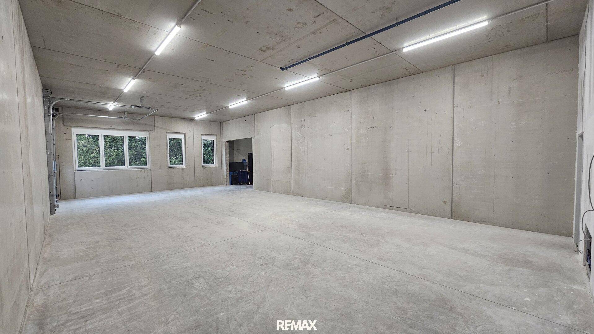 Heller, großer Raum mit Betonoberflächen, ausgestattet mit mehreren Fenstern und einem Zugang zu weiteren Bereichen.