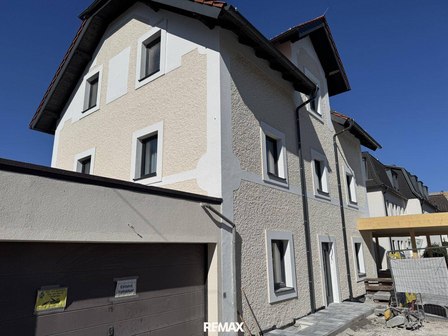 Seitenansicht eines Neubaus mit heller Fassade, dunklen Fenstern und einem Garagentor im Vordergrund.