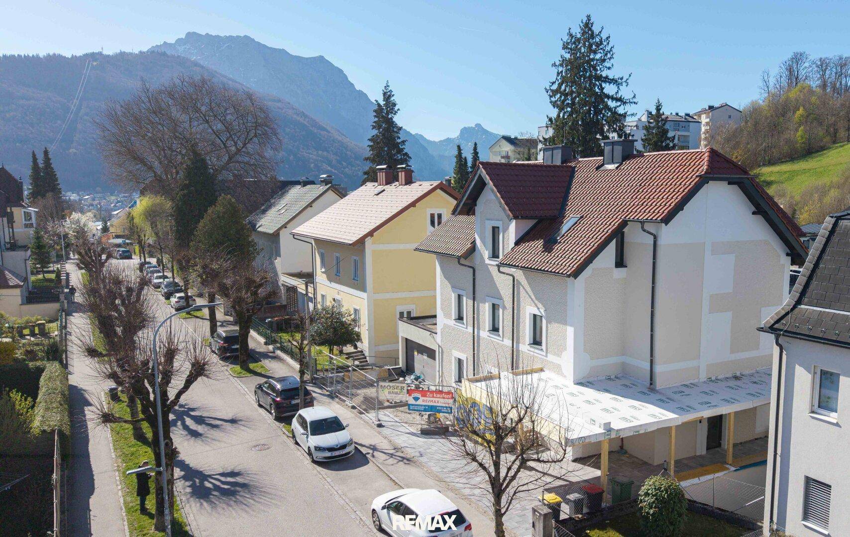 Blick auf eine Wohnstraße mit Häusern und Bergen im Hintergrund an einem sonnigen Tag.