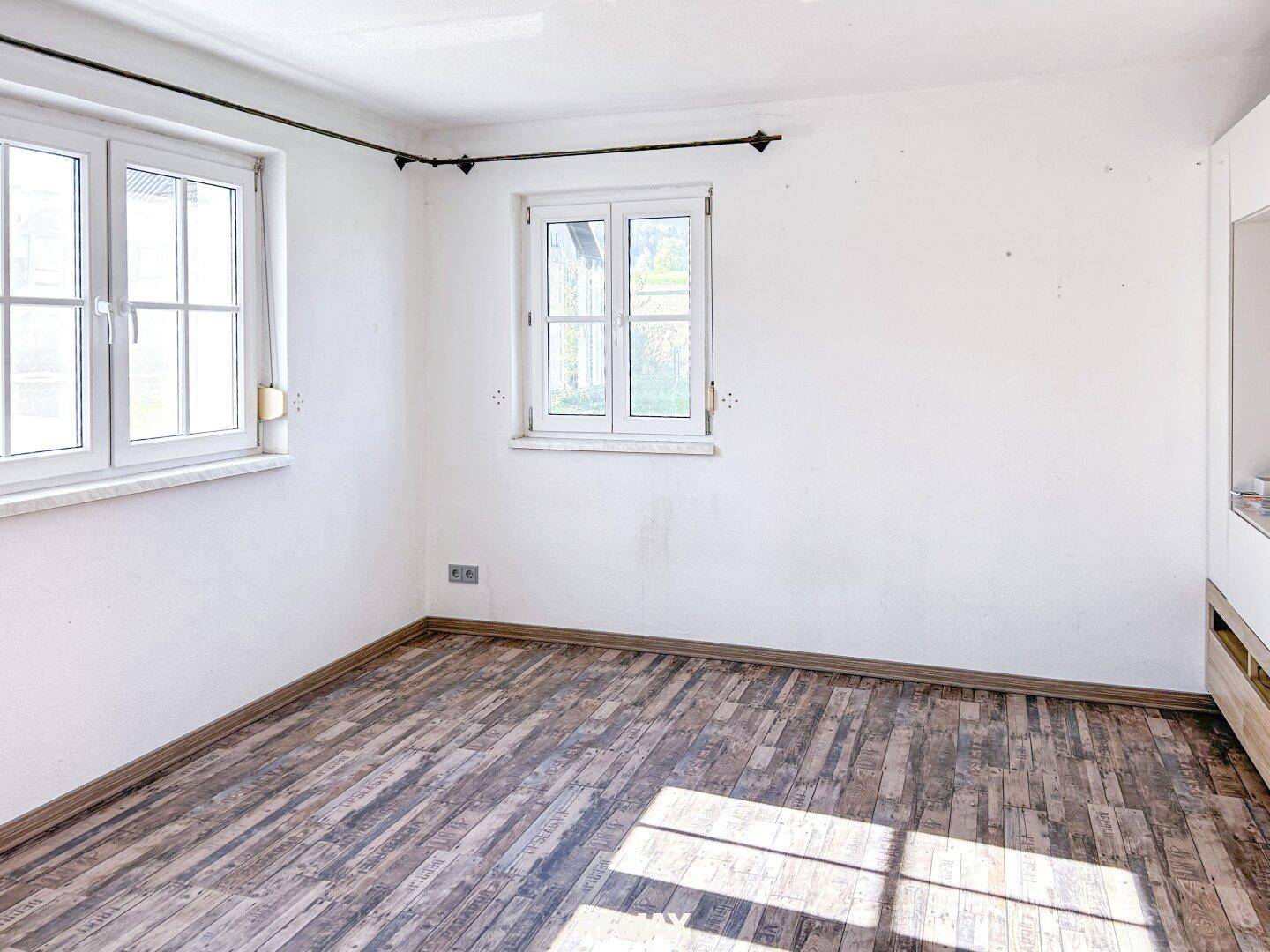 Heller, leerer Raum mit weißen Wänden und Holzbodenoptik, durch zwei Fenster gut belichtet.