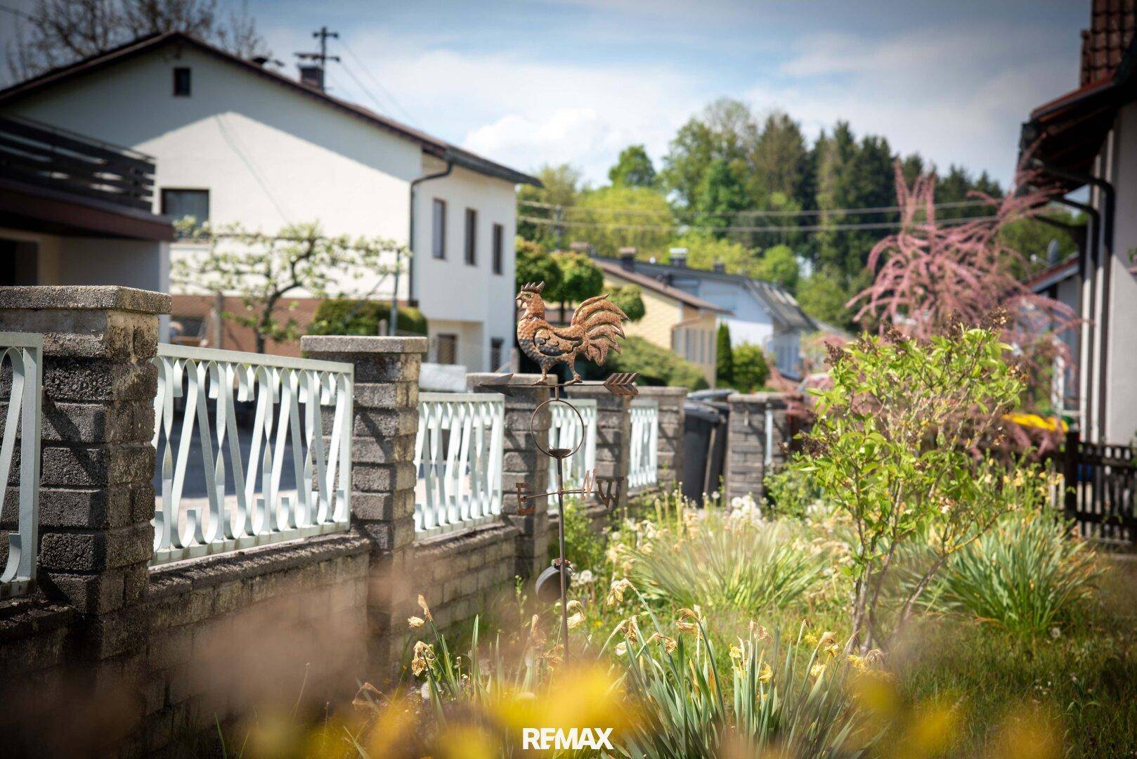 Detailansicht eines Gartenzauns mit dekorativem Wetterhahn und blühenden Pflanzen im Vordergrund.