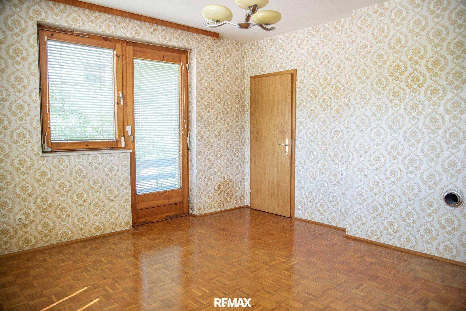 Leerer Raum mit gemusterten Tapeten, Parkettboden und Zugang zu einem Balkon.