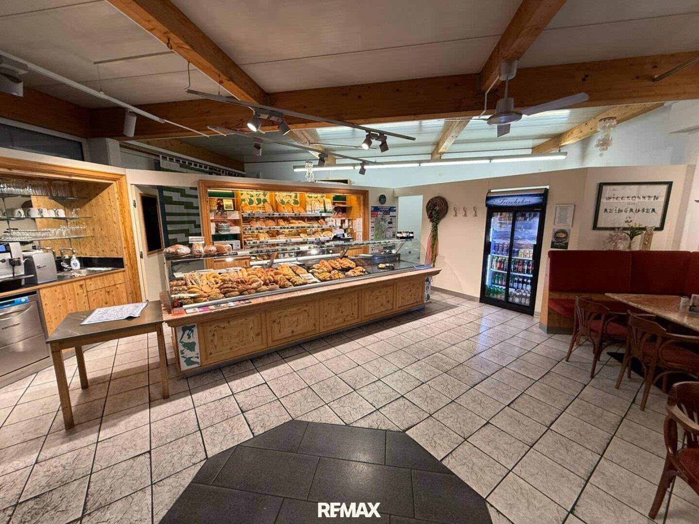 Innenansicht einer Bäckerei oder eines Cafés mit Verkaufstheke, Sitzgelegenheiten und Holzbalken.