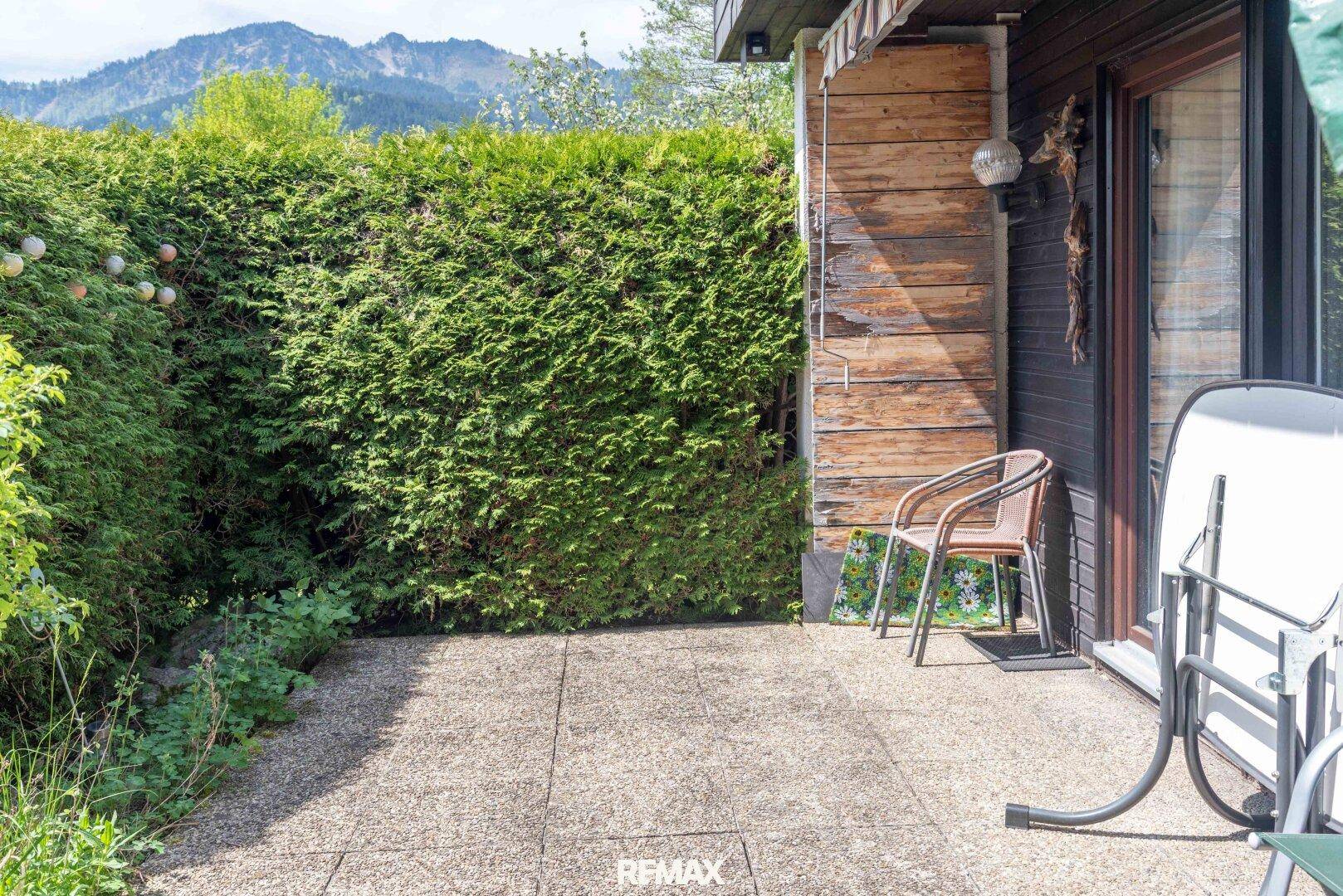 Kleine gepflasterte Terrasse mit hoher Hecke und Blick auf die umliegende Berglandschaft.