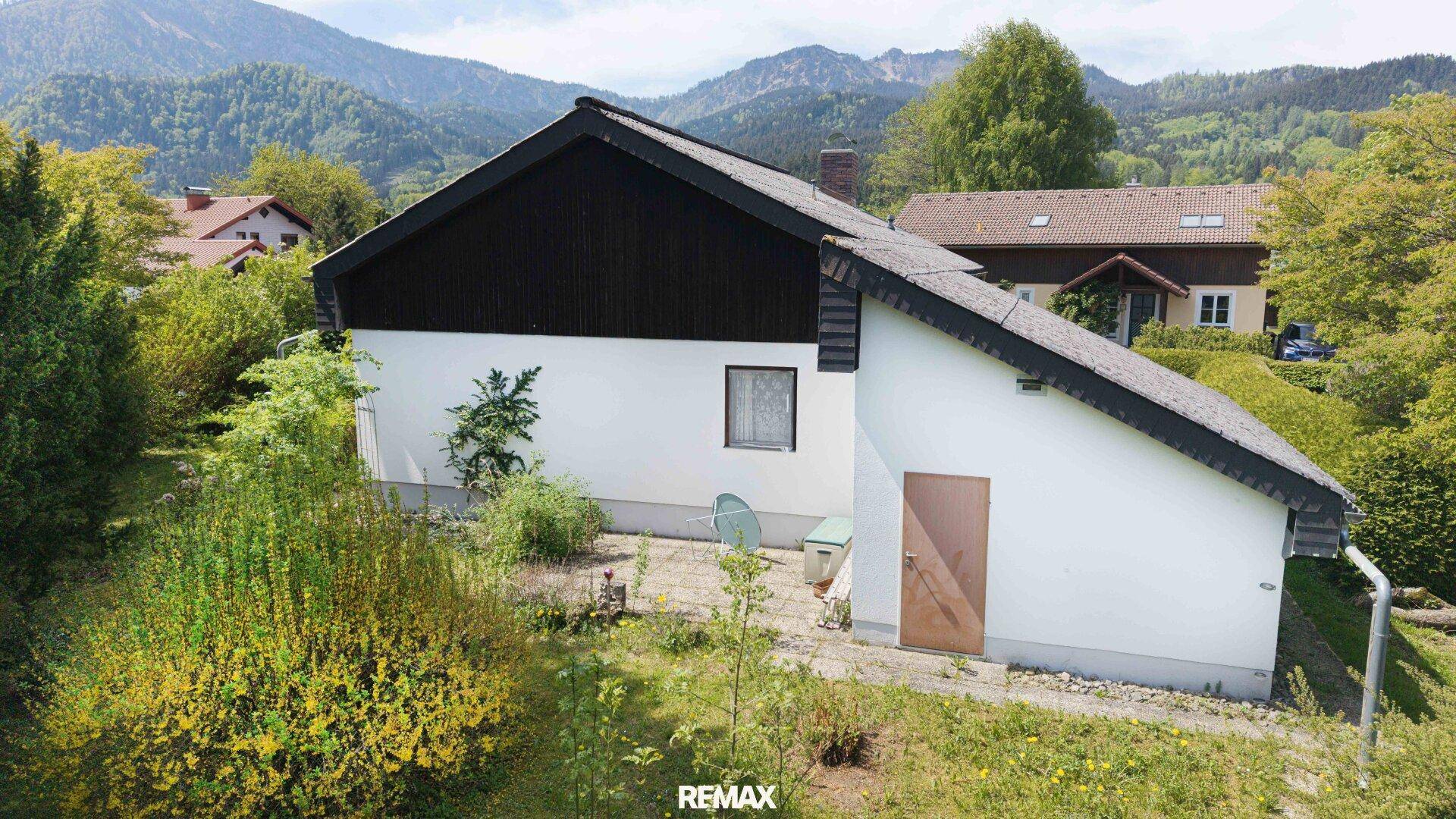Rückseite des Hauses mit weißer Fassade, dunklem Dach und einem naturbelassenen Gartenbereich.