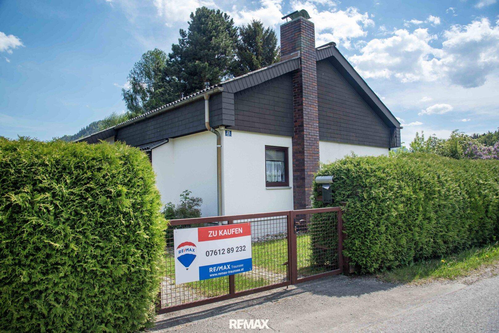 Hausansicht von der Straße mit einem Verkaufsplakat und einer hohen, gepflegten Hecke.