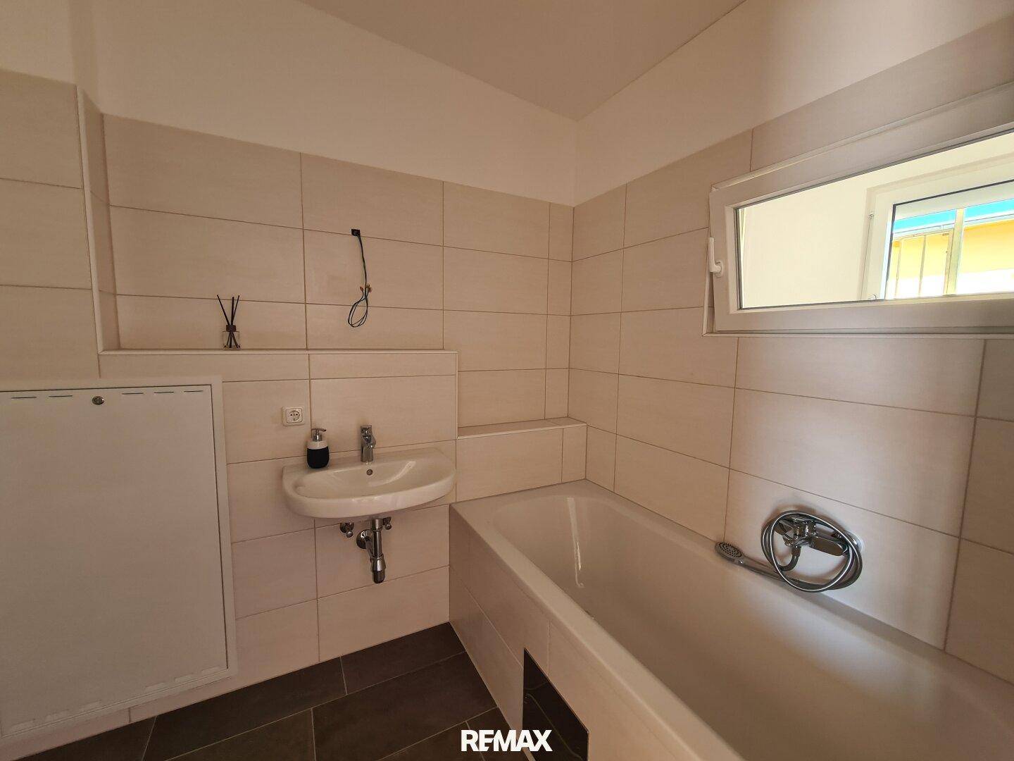 Funktionales Badezimmer mit beigefarbenen Fliesen, Badewanne, Waschbecken und einem Fenster für Tageslicht.