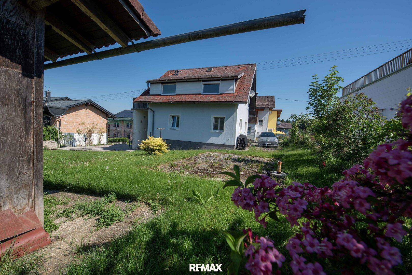 Blick auf ein Einfamilienhaus von einem grünen Gartenbereich mit blühenden Sträuchern im Vordergrund.