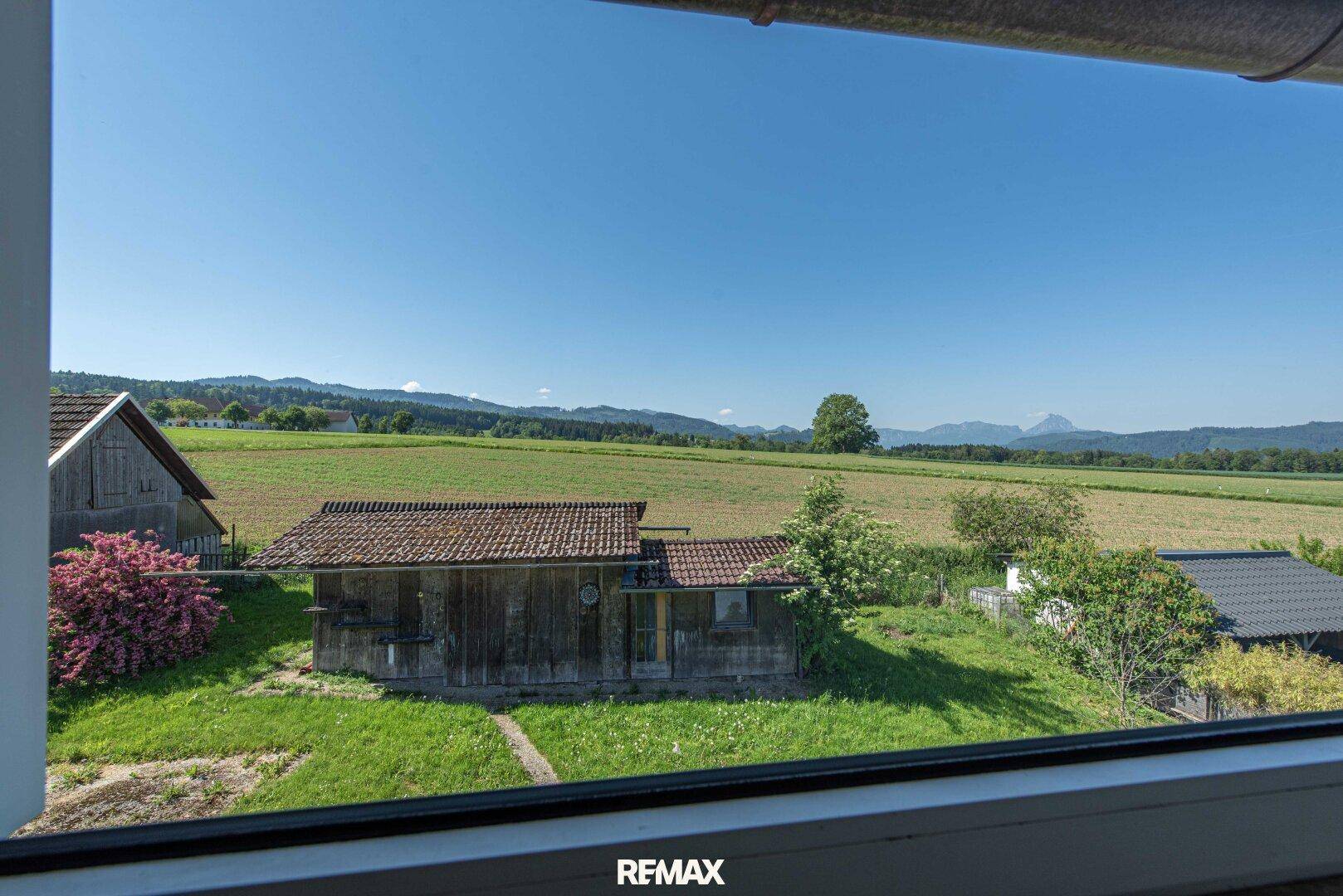 Weitläufiger Ausblick aus einem Fenster auf Felder, eine Holzhütte und ferne Berge unter blauem Himmel.