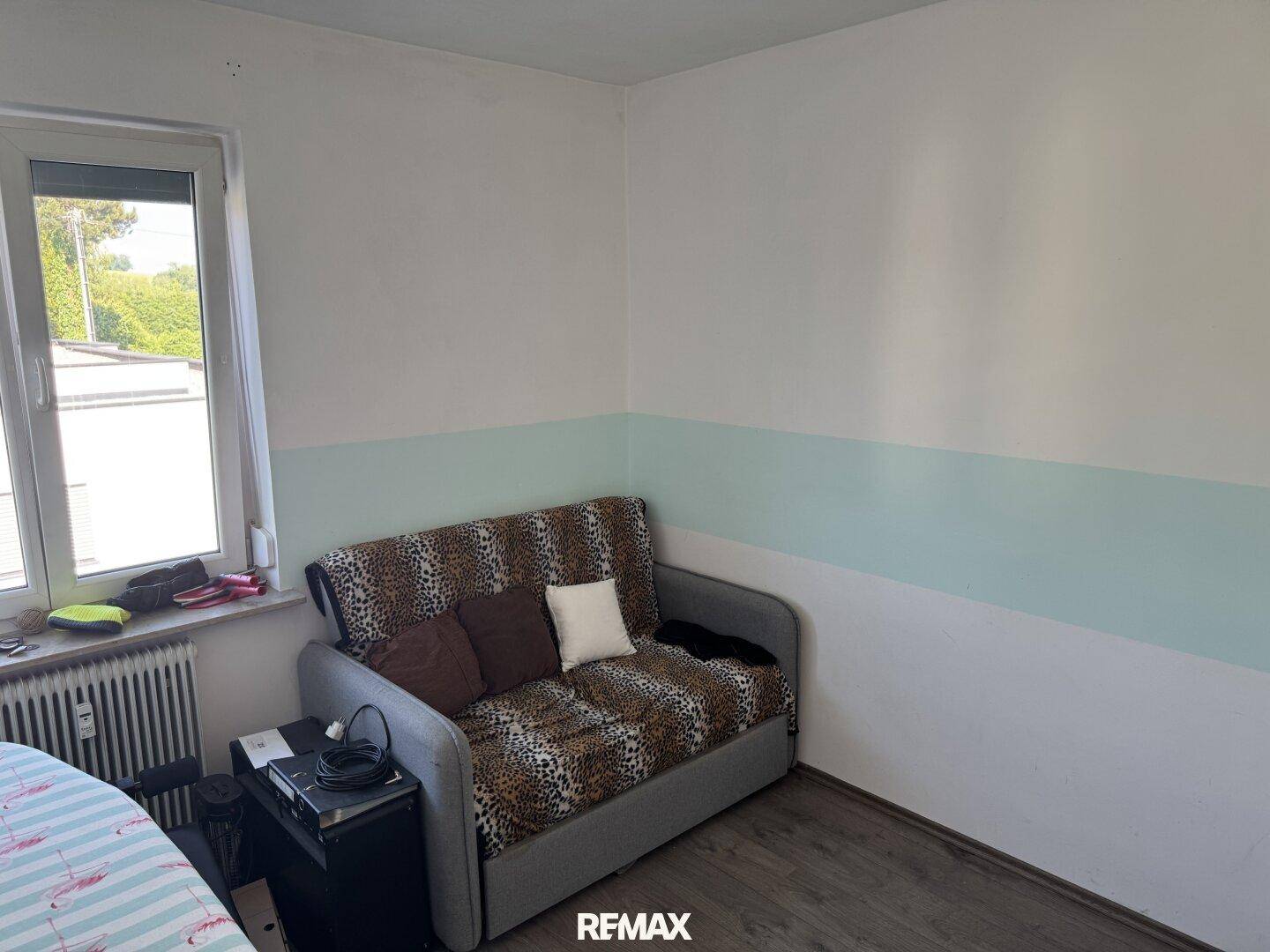 Kleines Schlafzimmer mit Schlafsofa, Laminatboden und einem Fenster mit Blick nach draußen.