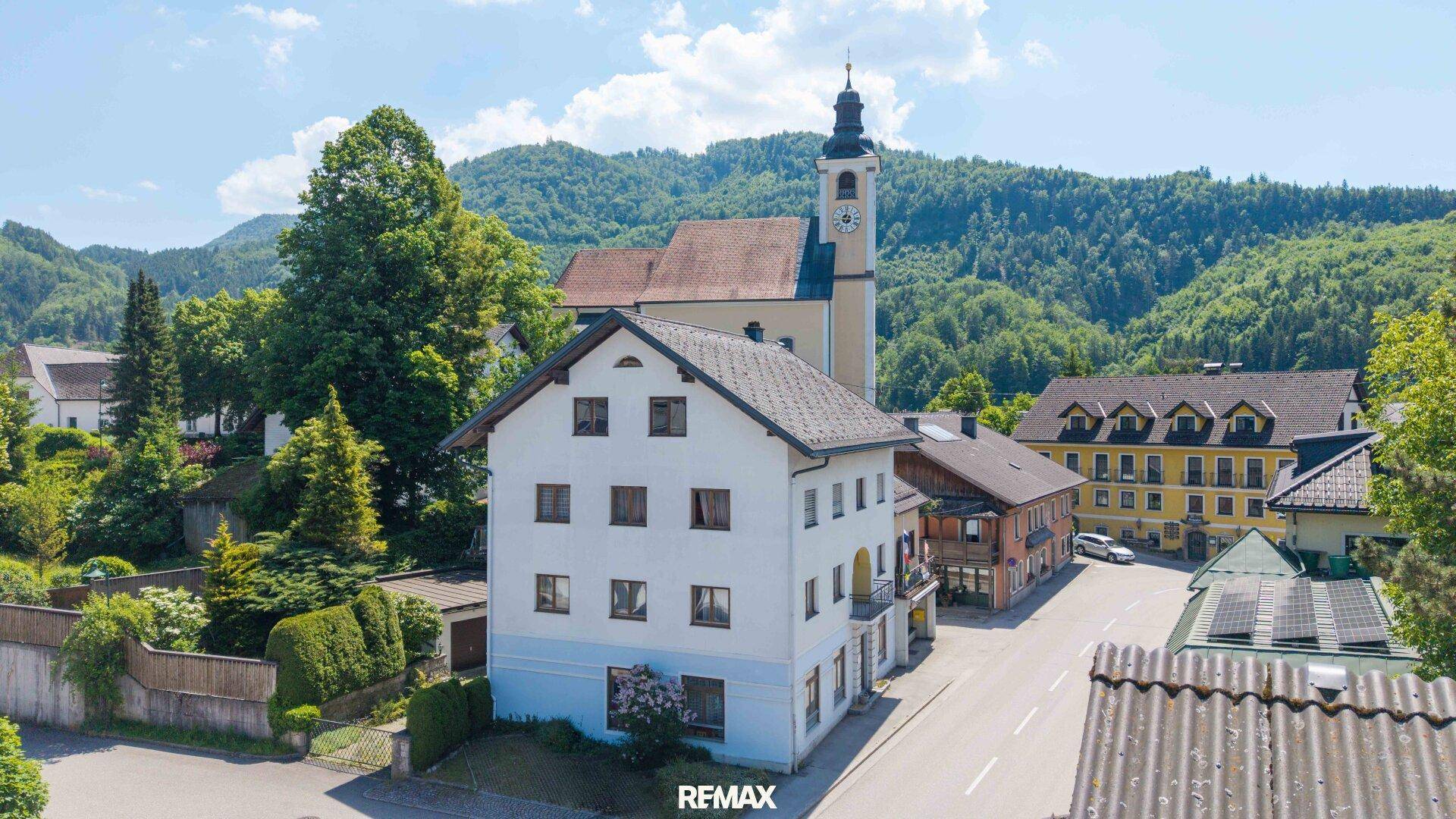 Luftaufnahme des Gebäudes mit umliegender Landschaft und Kirche im Hintergrund.