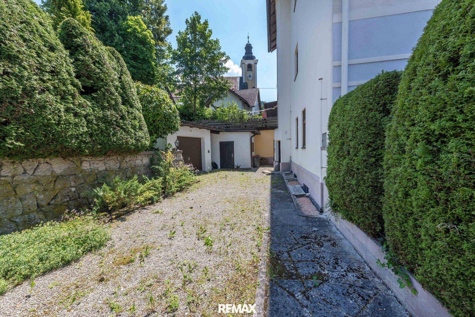 Schotterweg führt zu einer Garage neben dem Haus, umgeben von hohen Hecken und einem Kirchturm im Hintergrund.