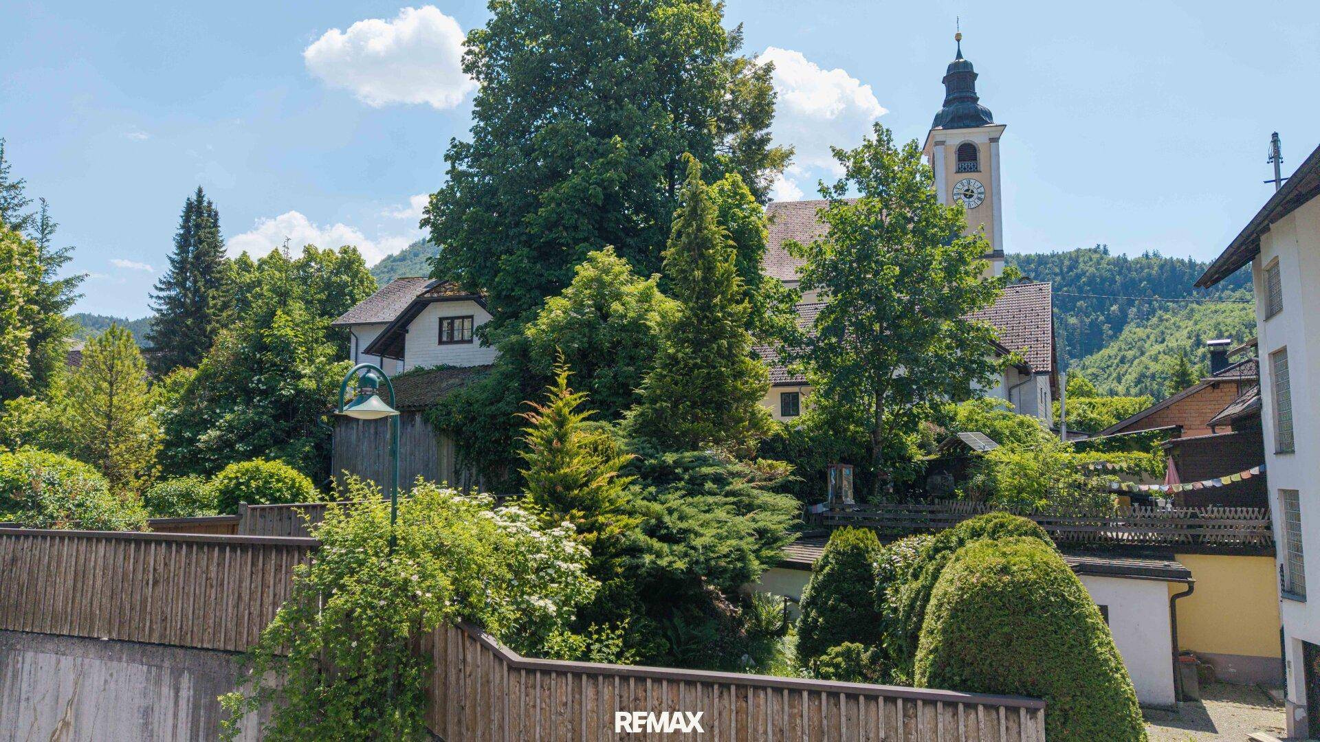 Malerische Umgebung mit einem Kirchturm, üppigem Grün und traditionellen Häusern unter blauem Himmel.