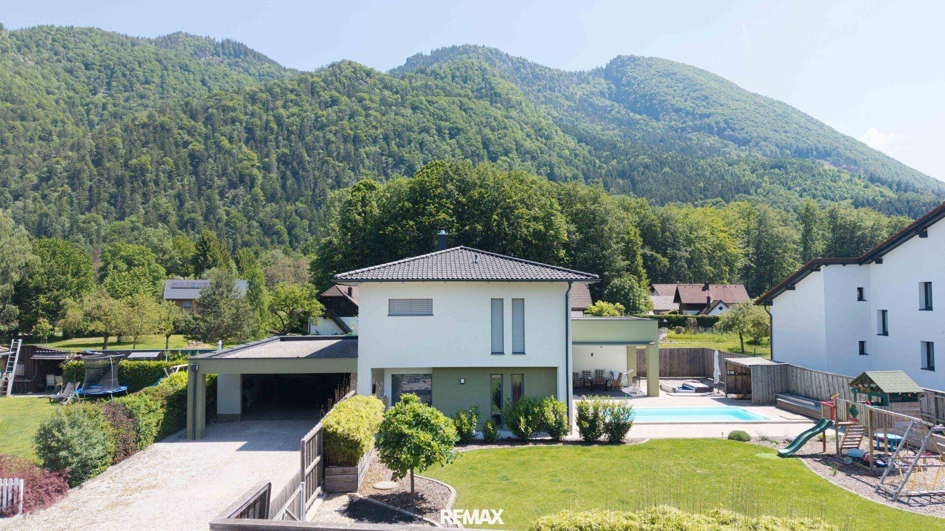 Luftaufnahme eines modernen Hauses mit großem Garten, Pool und Carport, eingebettet in eine grüne Berglandschaft.