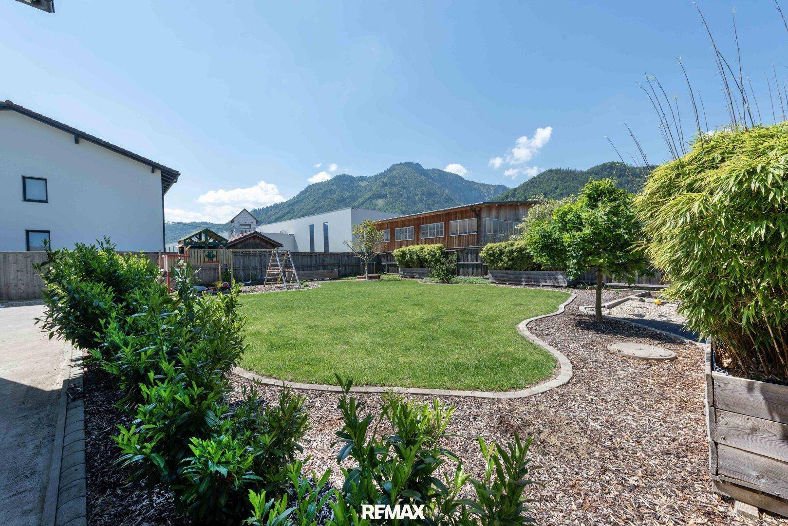 Weitläufiger Gartenbereich mit Rasen, Sträuchern und einem Spielplatz, umgeben von einer ruhigen Berglandschaft.