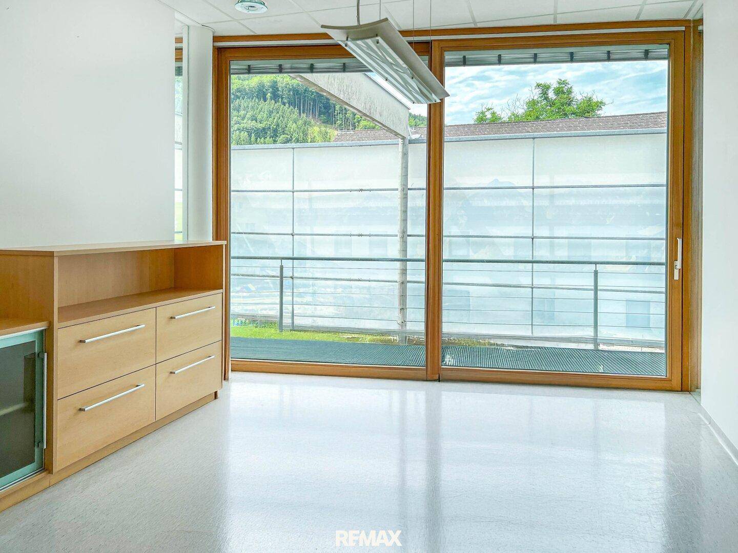 Heller Büroraum mit großen Holzfenstern und Blick auf grüne Hügel und einen Balkon.