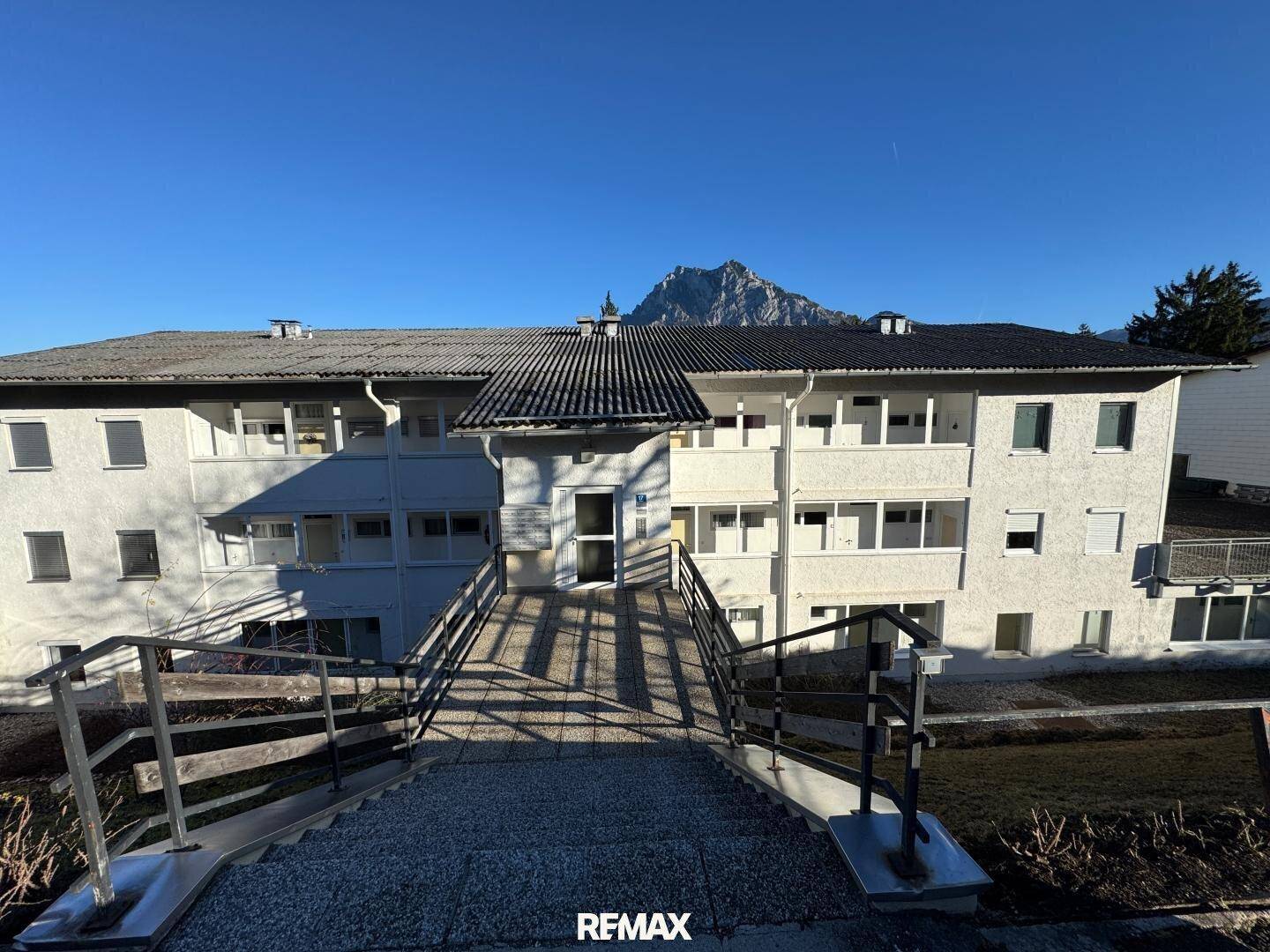 Mehrfamilienhaus mit Zugang über eine Rampe und Bergblick im Hintergrund.