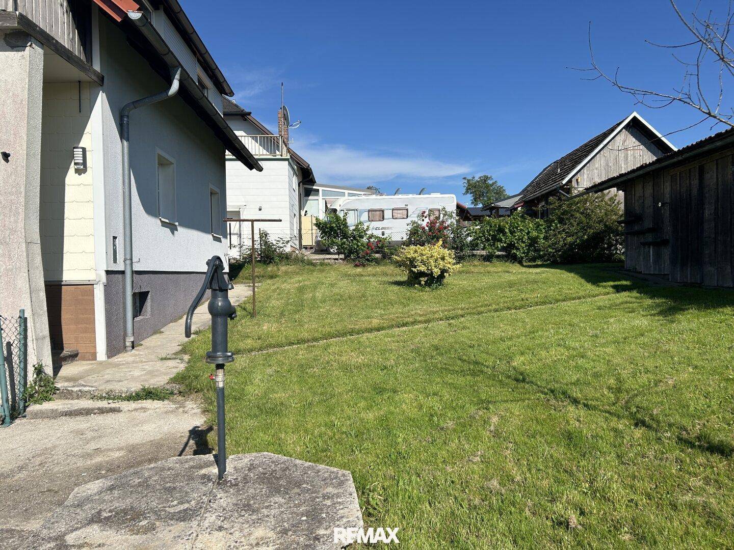 Grüner Gartenbereich neben dem Haus mit einer alten Handschwengelpumpe und einem Wohnwagen im Hintergrund.
