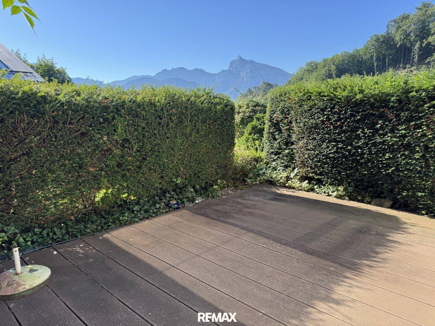 Holzterrasse mit Hecken und Blick auf die umliegende grüne Landschaft und Berge.