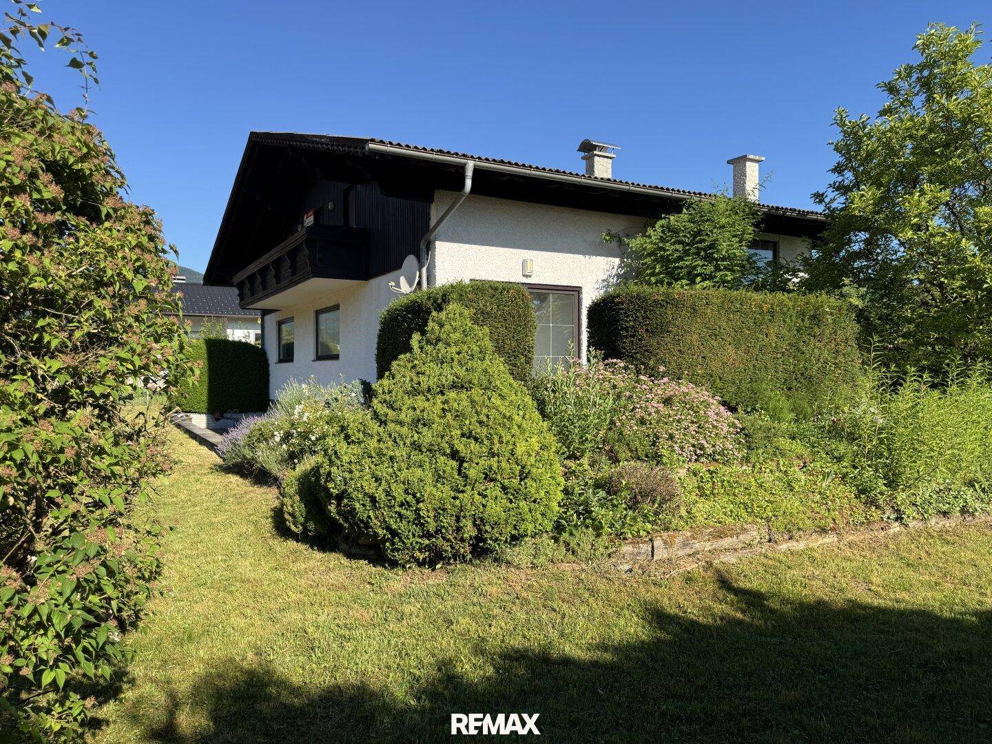Seitlicher Blick auf das Haus mit gepflegtem Garten, Hecken und blühenden Sträuchern unter blauem Himmel.