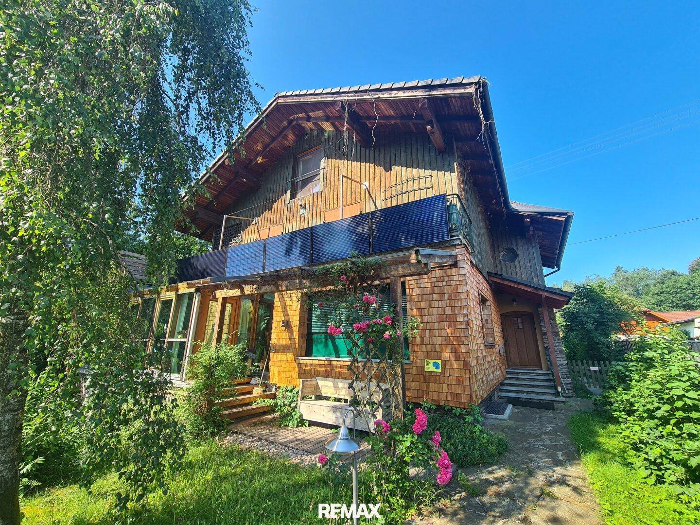 Rustikale Holzschindelfassade des Hauses mit Balkon und üppiger grüner Bepflanzung.