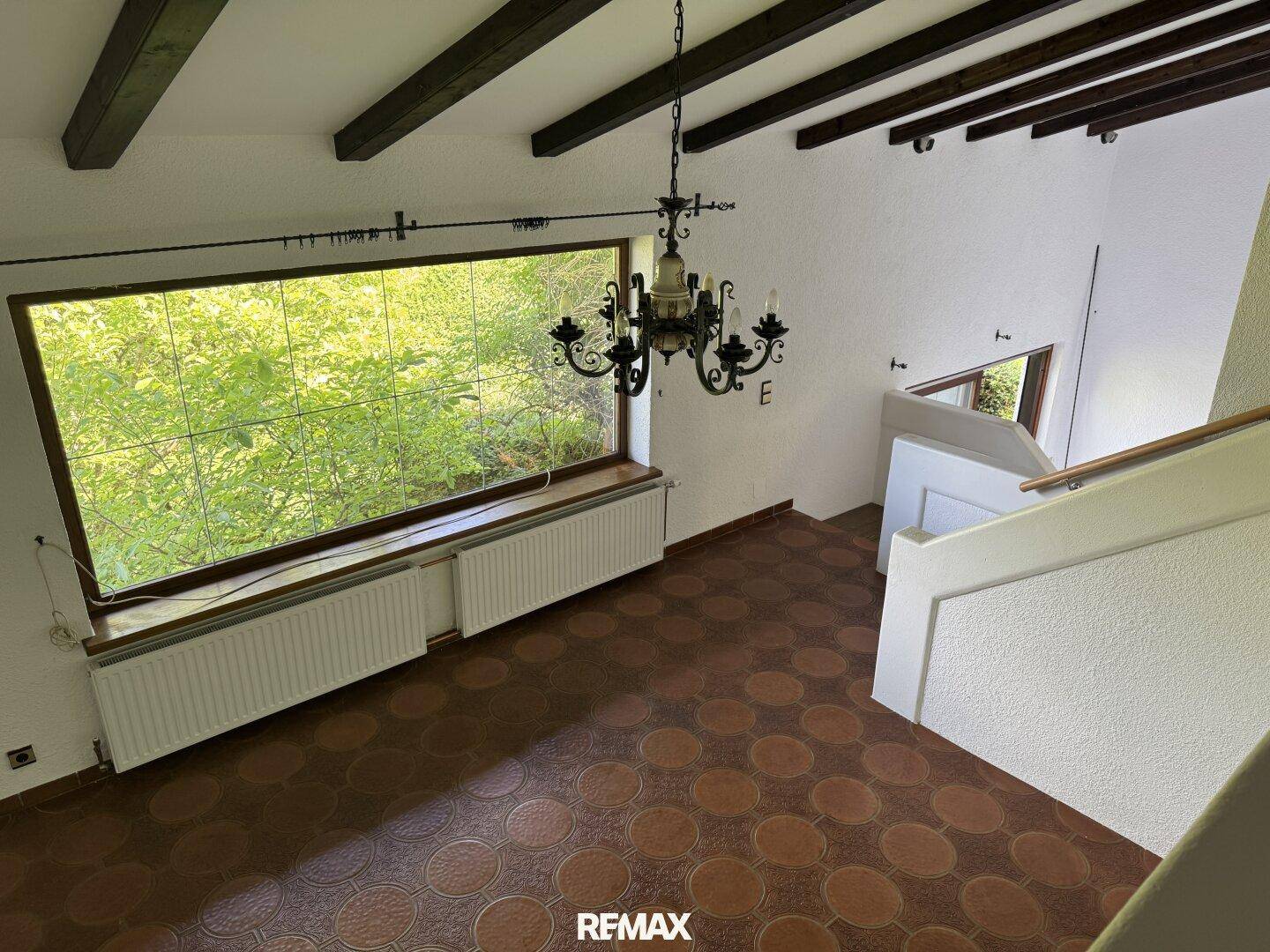 Geräumiger Wohnbereich mit hohen Decken, Holzbalken und einem großen Fenster mit Blick ins Grüne.